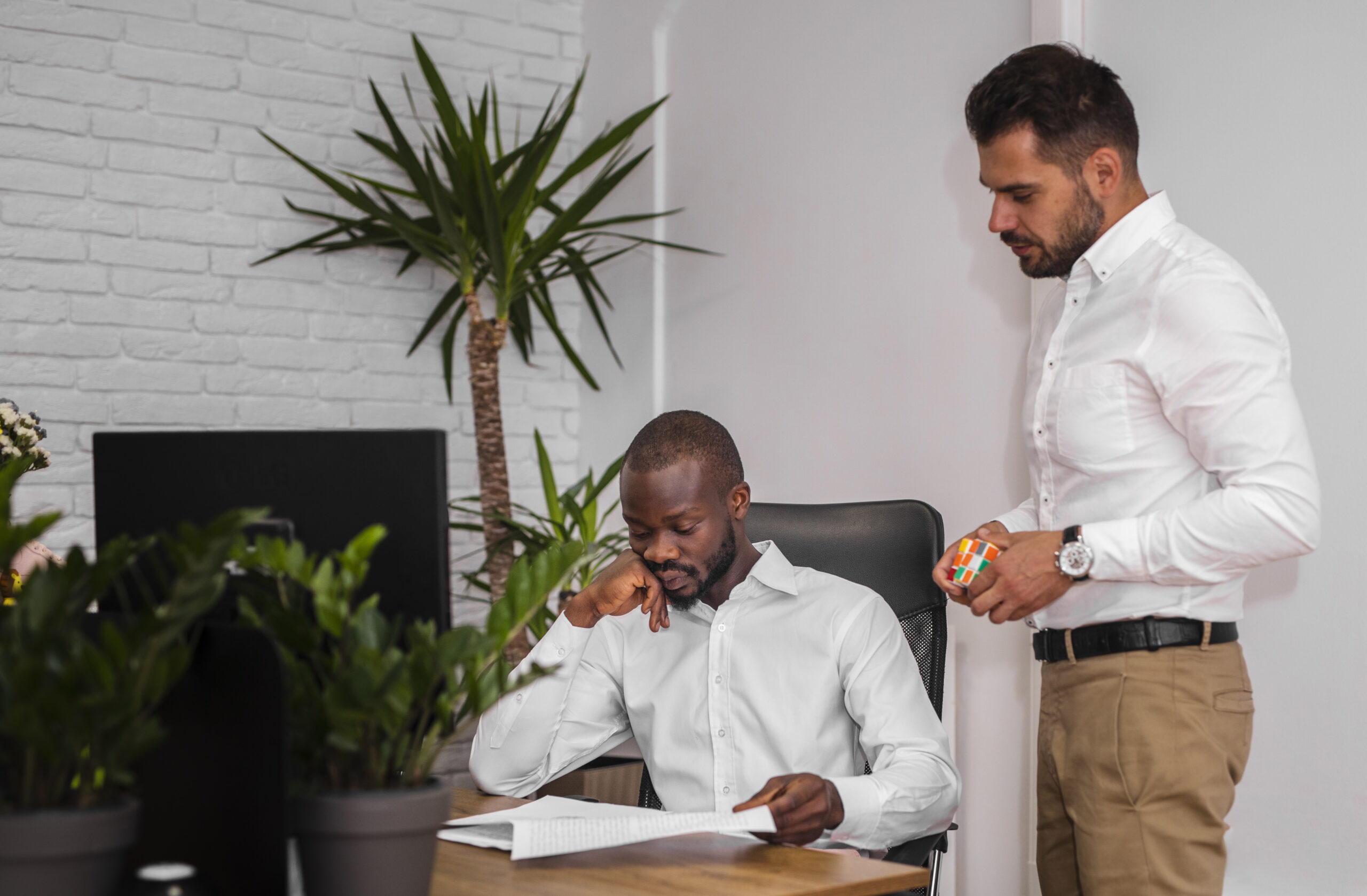 Two men in an office, one sitting at a desk with documents, the other standing and holding a Rubik's cube